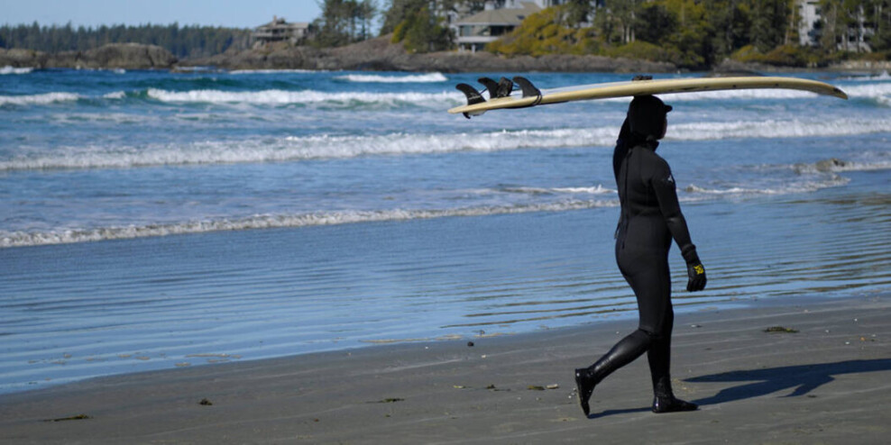 A female surfer in a wet suit walks on the shore carrying a surfboard over her head.