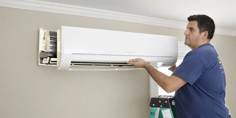 A man in a blue t-shirt installs a heat pump on a beige wall.