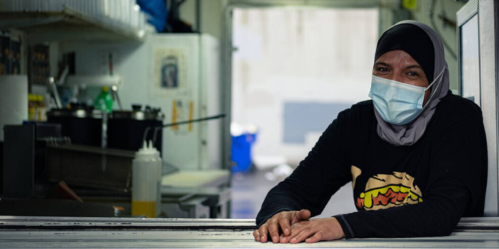 A woman wearing a head scarf and a mask sits in a commercial kitchen.