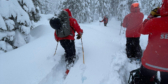 Search and rescue volunteers in red coats and black snowpants trudge through knee deep snow.