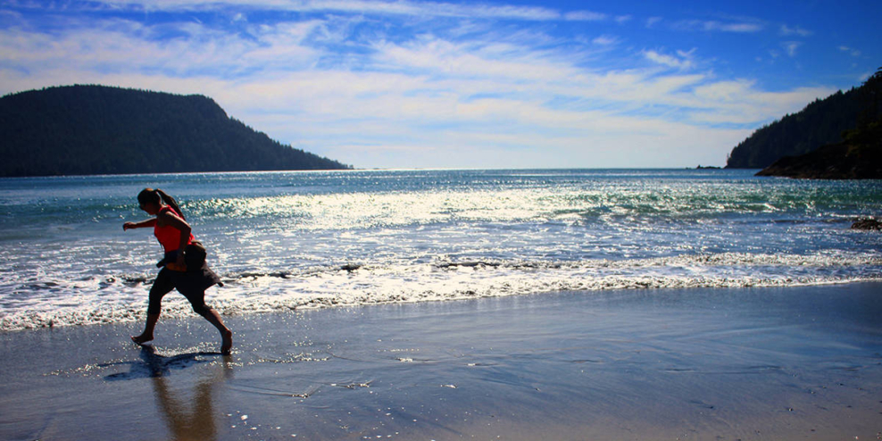 A woman puts her feet in the ocean on a sunny day. There are mountains in the background.